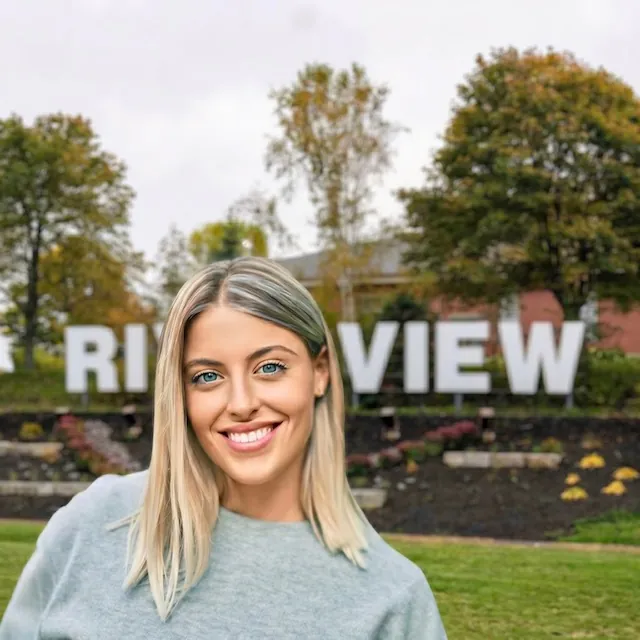 Smiling blonde woman with bright white teeth in front of the Riverview NB sign after a teeth cleaning at Bright White Dental Hygiene Clinic